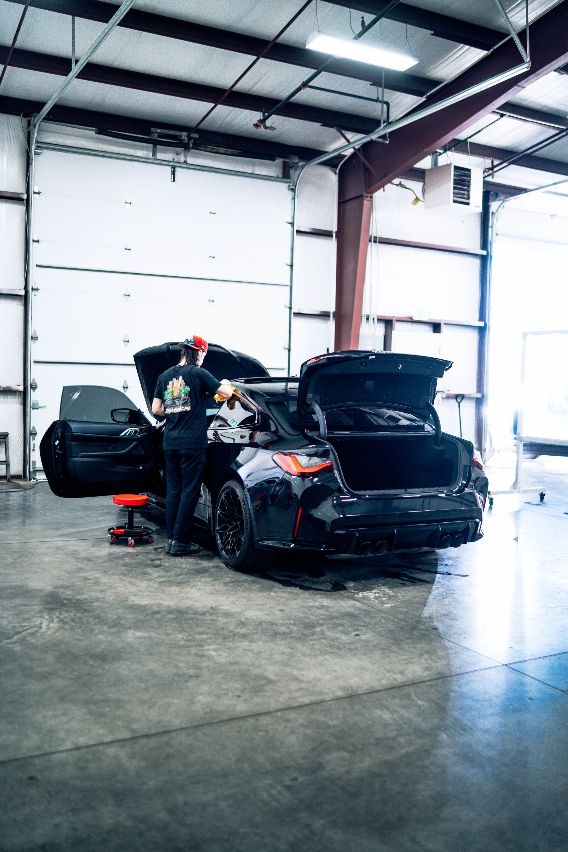 Two black cars with open trunks in a garage; one person works between them.