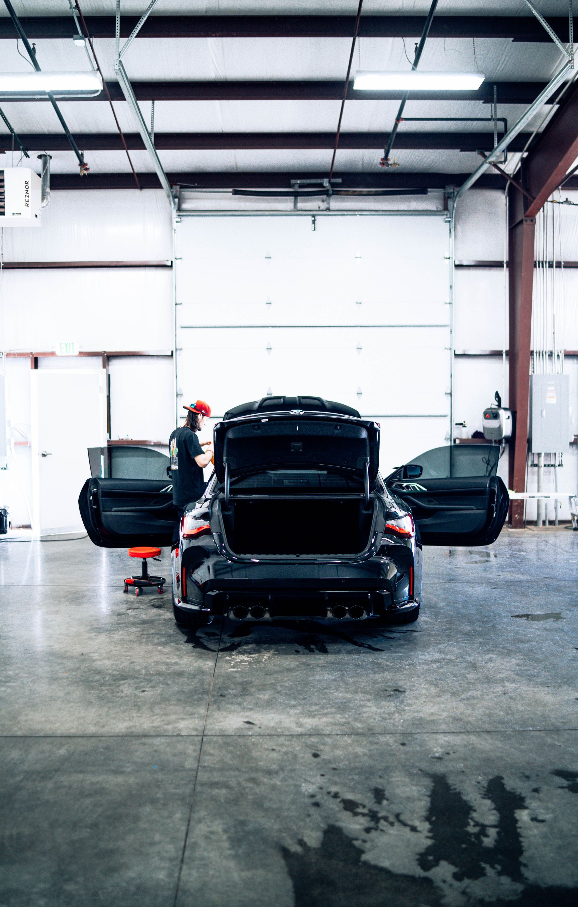 Black car in a large auto shop with trunk and doors open, mechanic nearby.