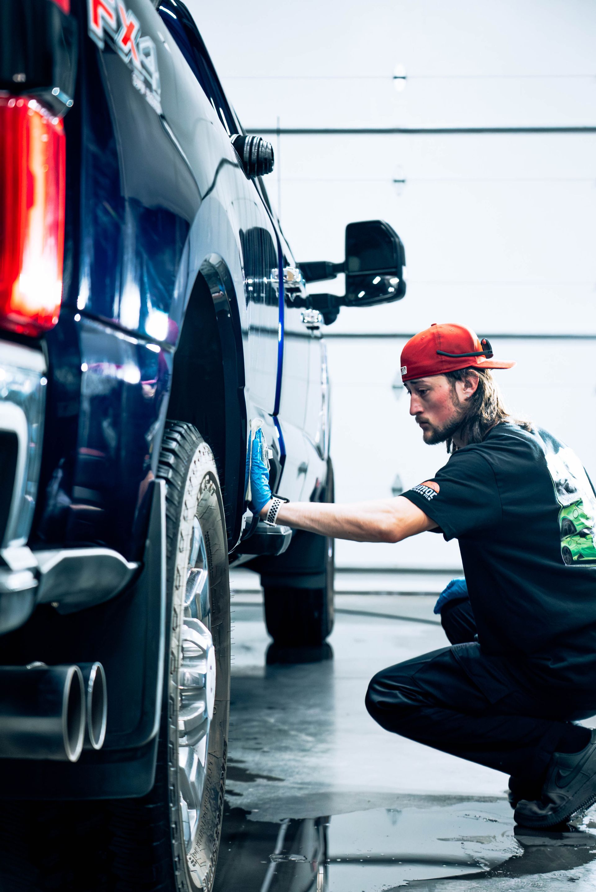 Mechanic in red cap inspecting the rear wheel of a blue truck in a garage.