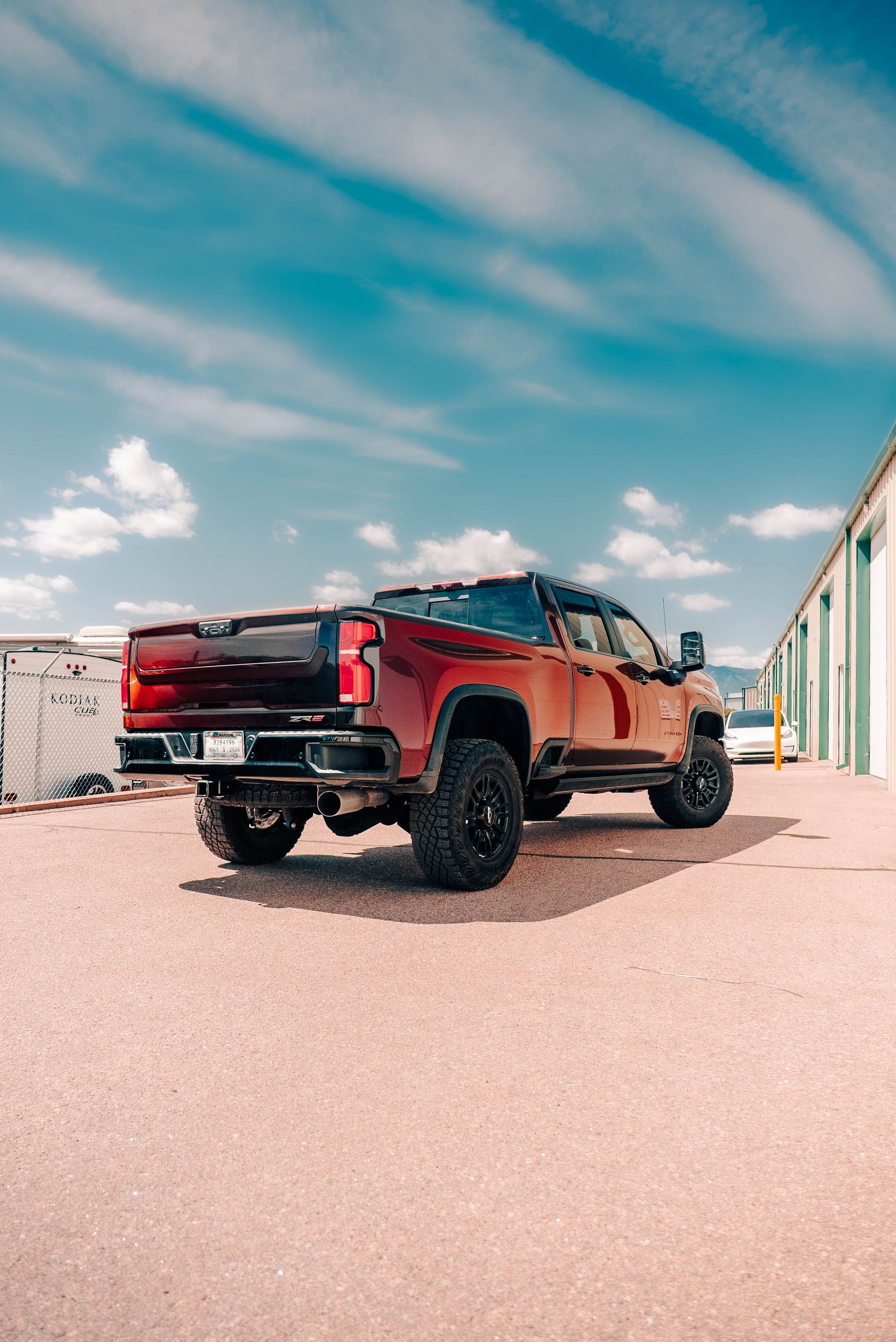 Red lifted pickup truck parked on gravel beside industrial buildings under a blue sky.