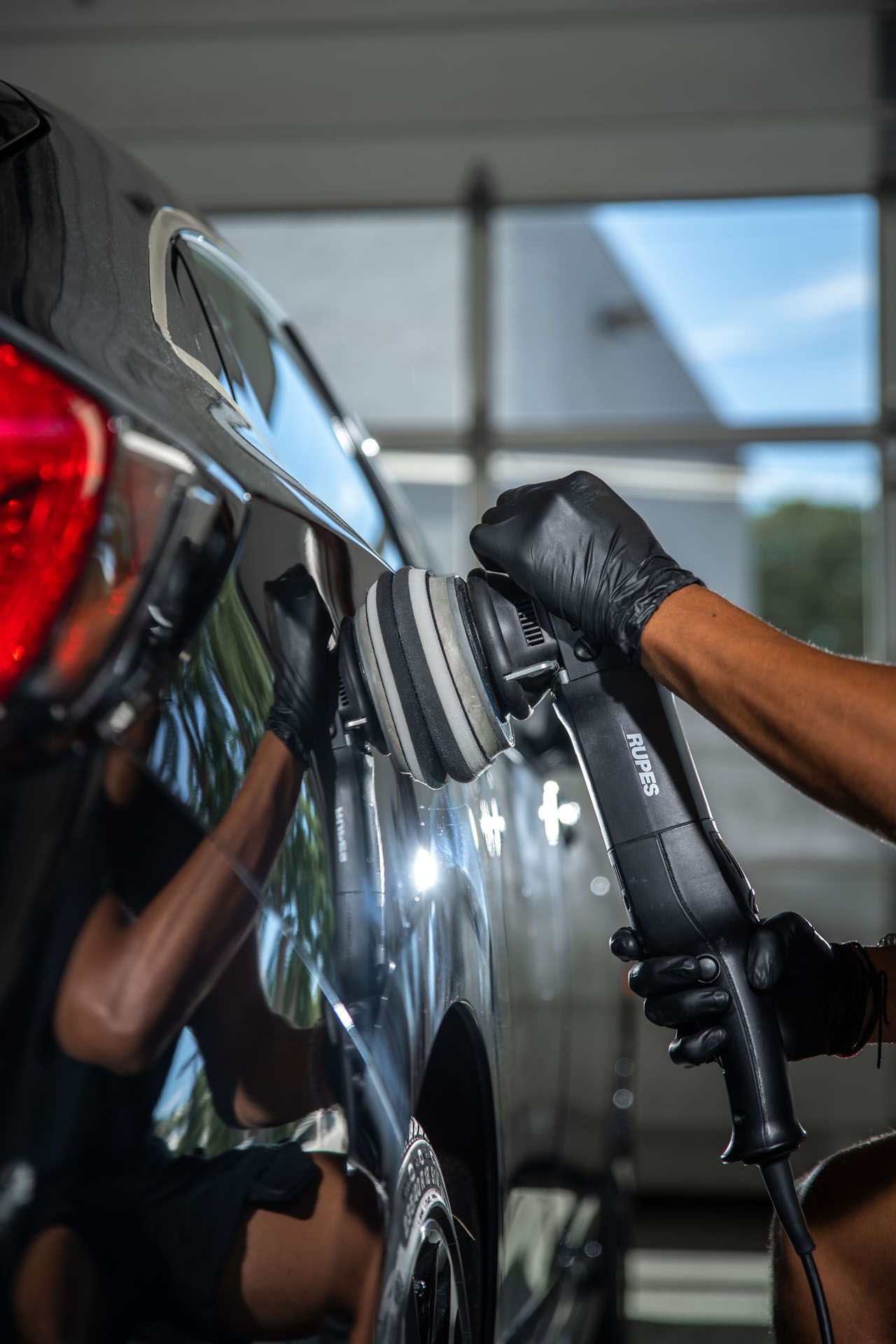 Gloved hand polishing a black car with a buffer in a garage