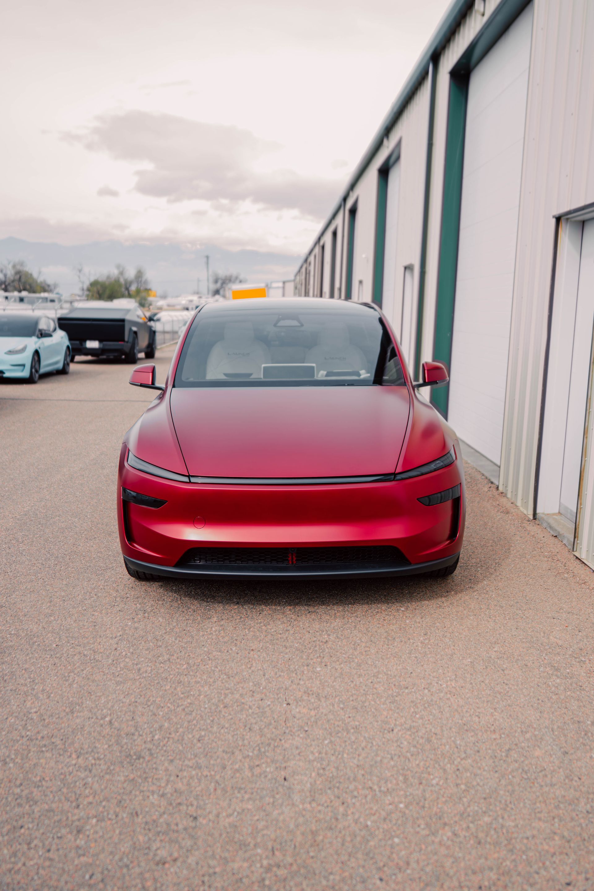 Red Tesla parked beside a warehouse on a gravel lot under an overcast sky