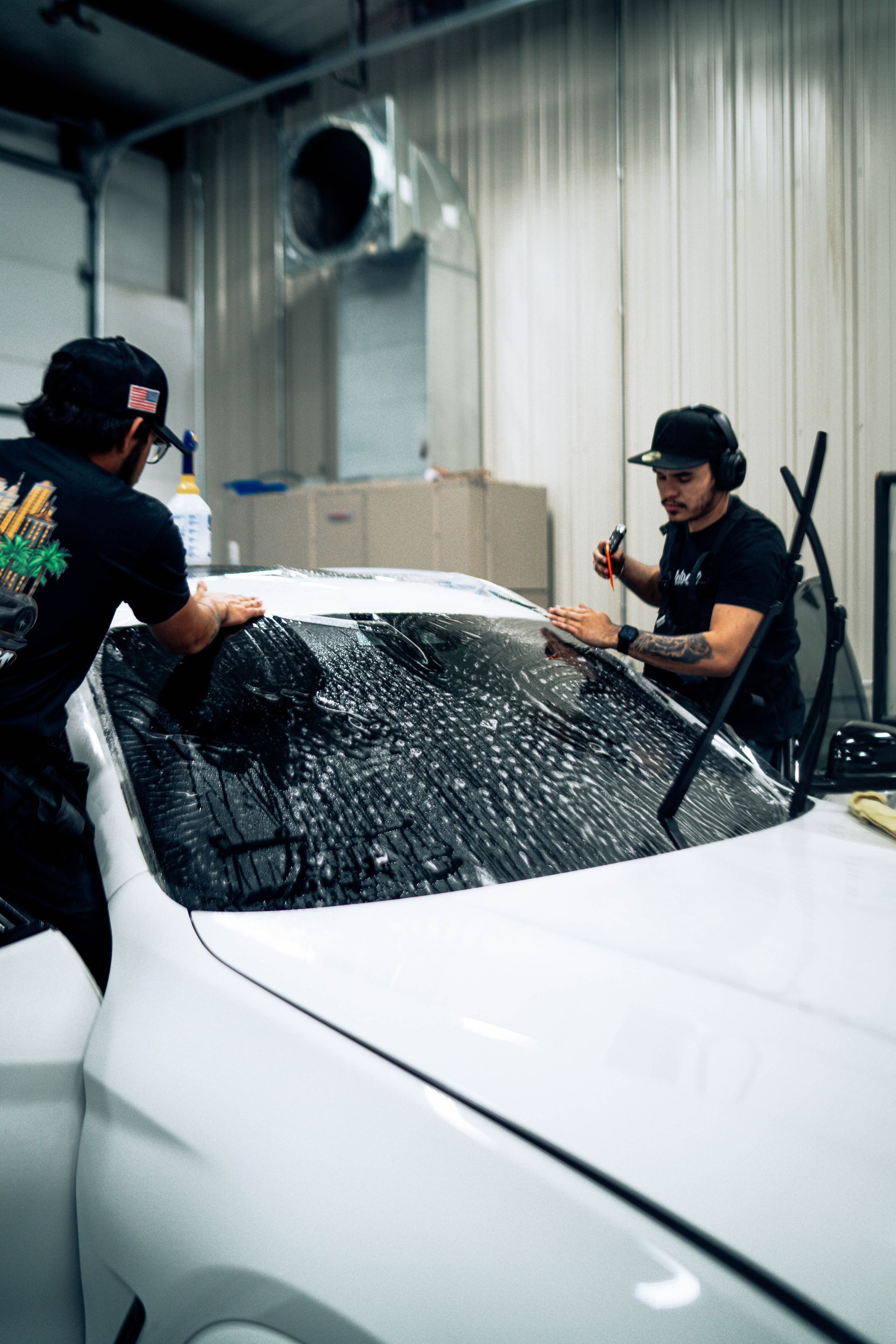 Two workers washing a white car with hoses in an indoor garage, covered in soap and water splashes