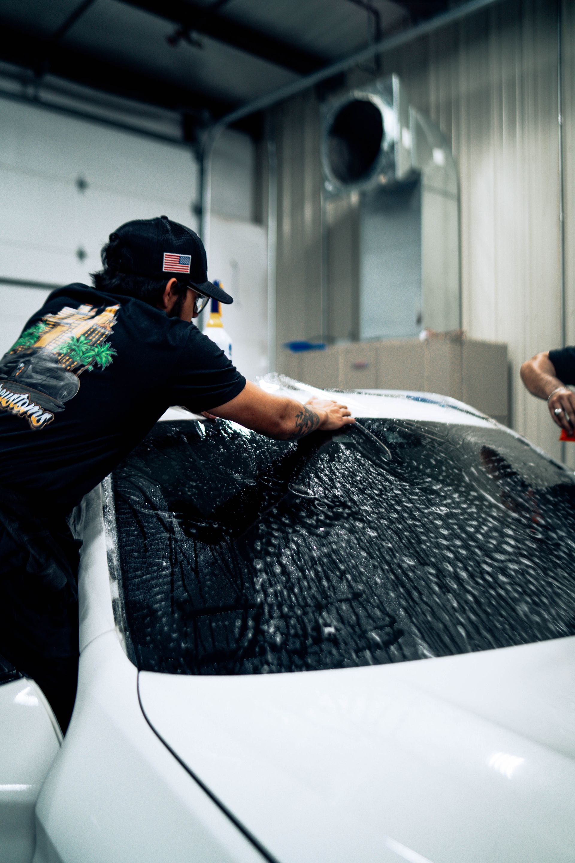 Person washing a white car in a garage, spraying water over the windshield