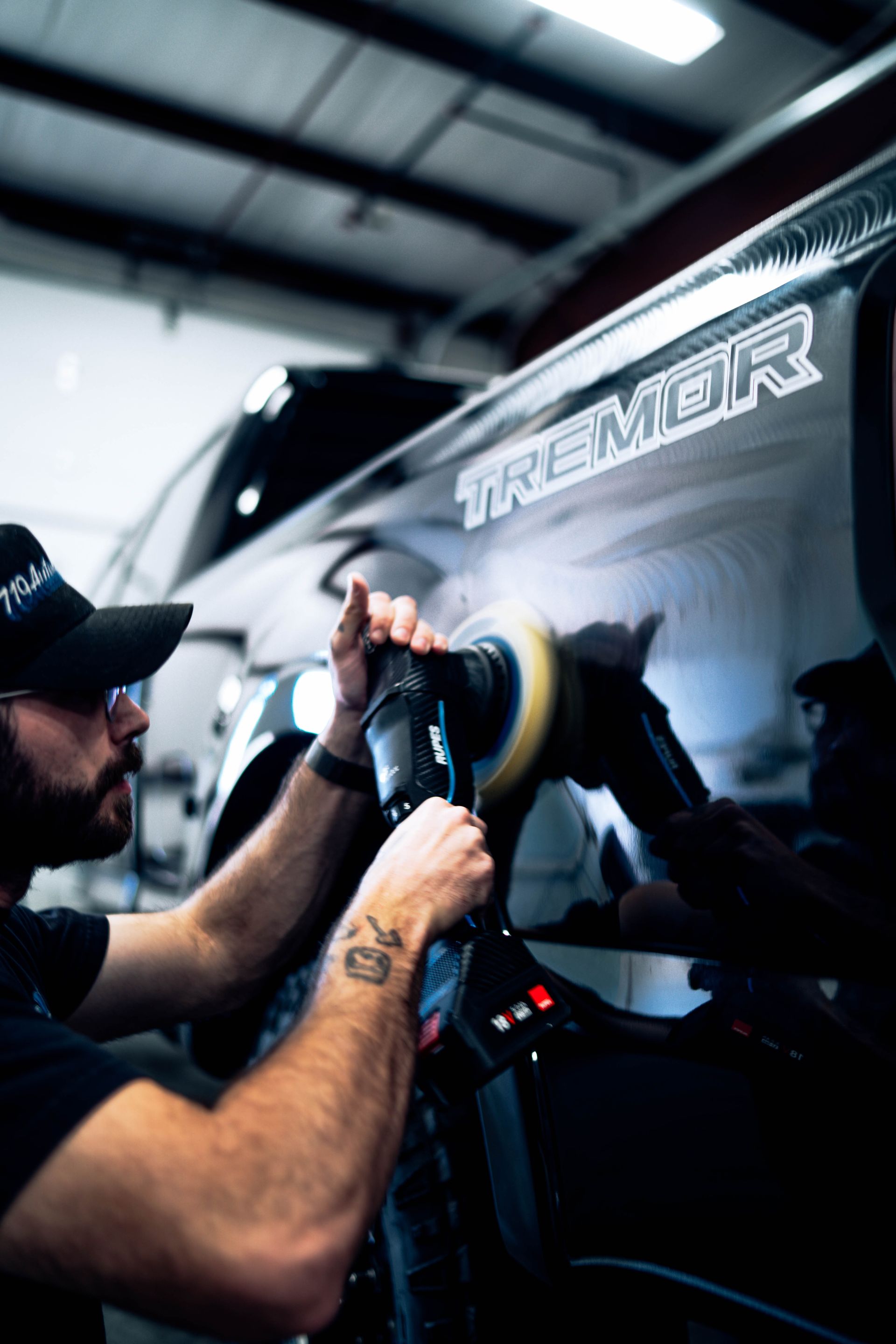 Man polishing a black car panel in a garage, using a buffing tool under bright workshop lights