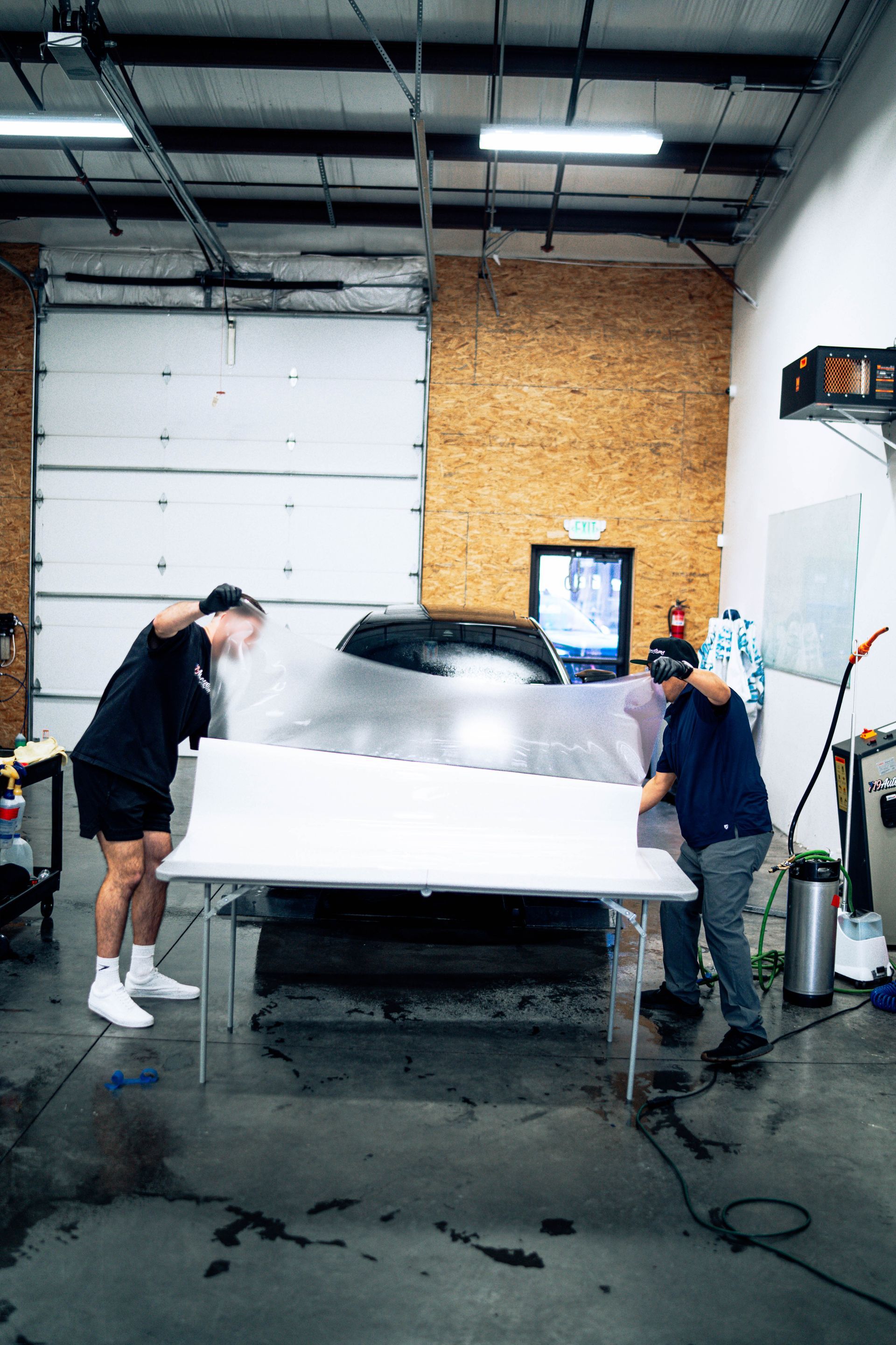 Two people hold a white panel in a garage workshop with a car wrapped in plastic behind them.