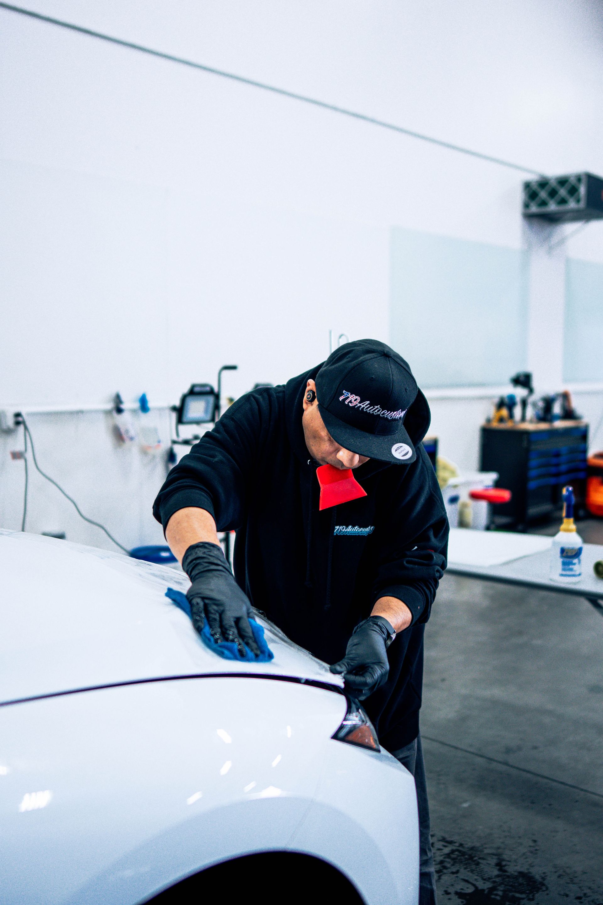 Person in black cap and gloves polishing a white car in a workshop