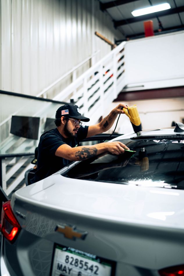 Worker polishing a white car in a garage, with stairs and overhead lights in the background