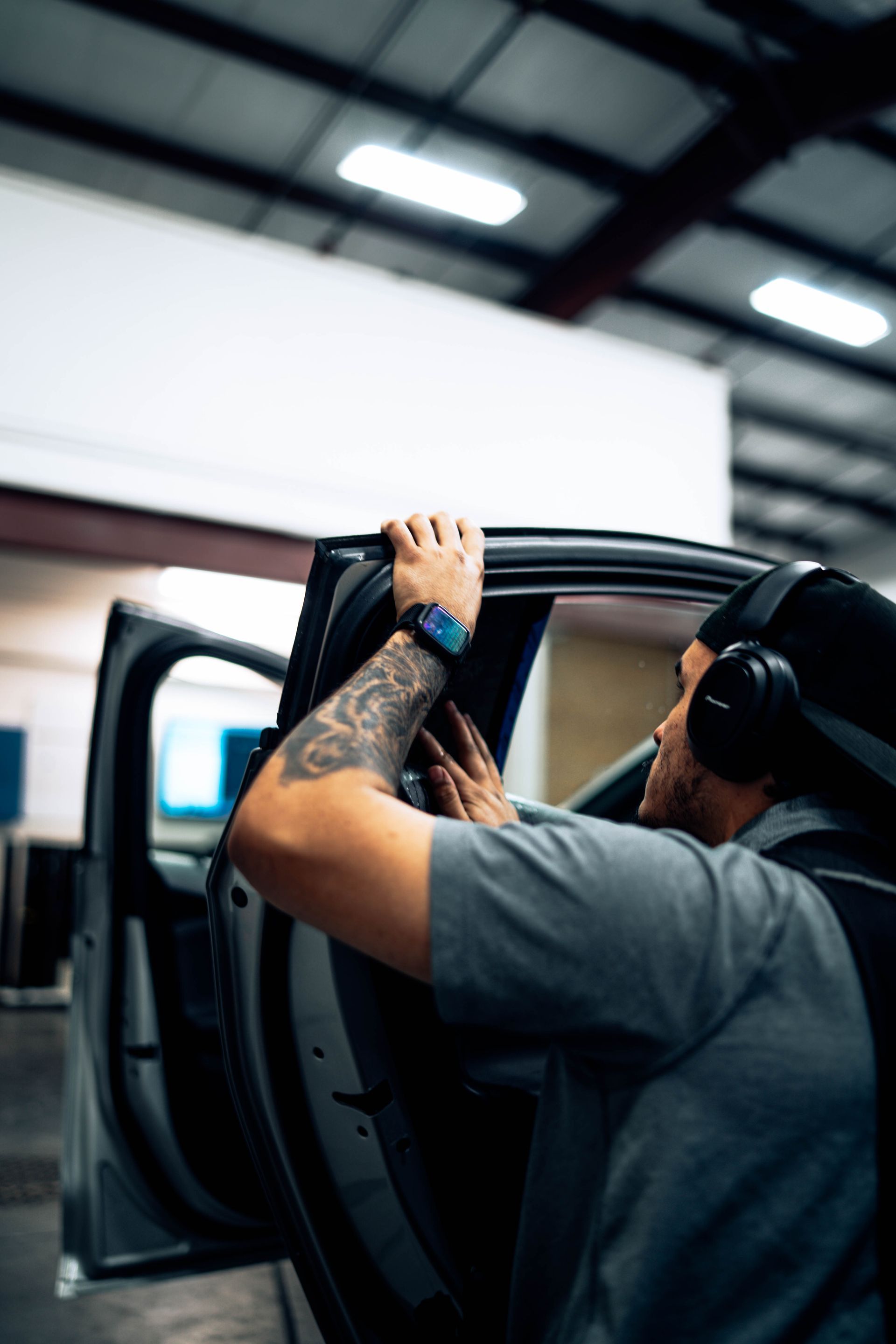 Person loading a black car door in an auto shop, wearing headphones and a tattooed arm visible