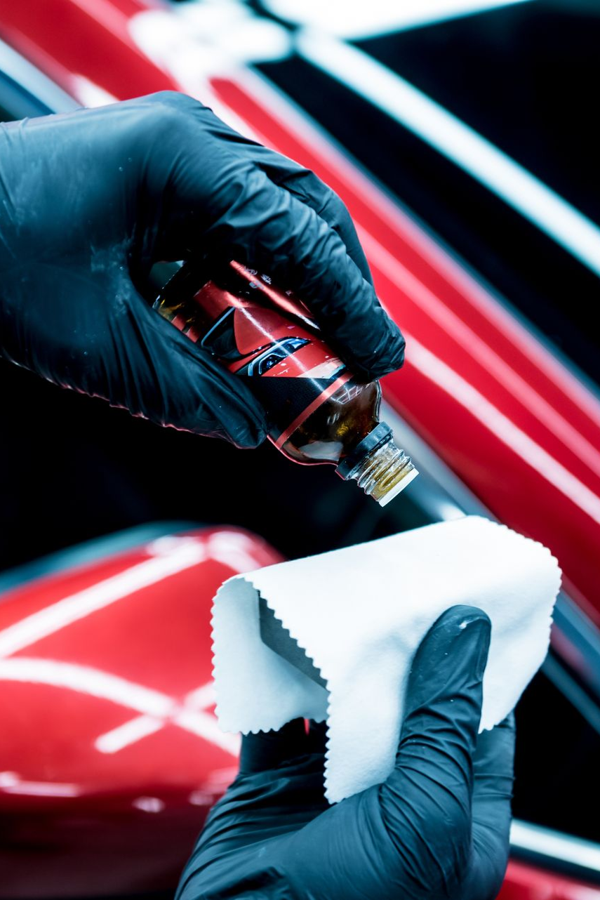 A person wearing black nitrile gloves applies a ceramic coating liquid from a bottle onto a white applicator cloth.