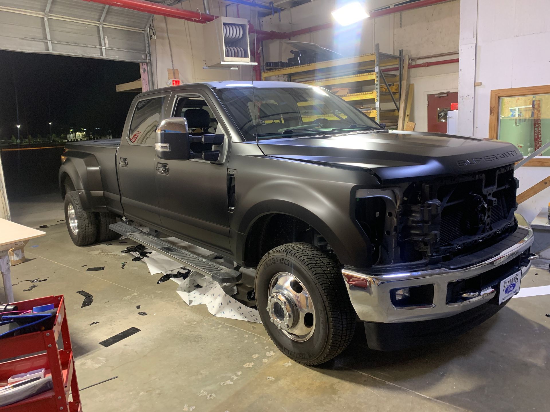A matte black dually pickup truck parked inside a workshop with its front grille and headlights removed for service.