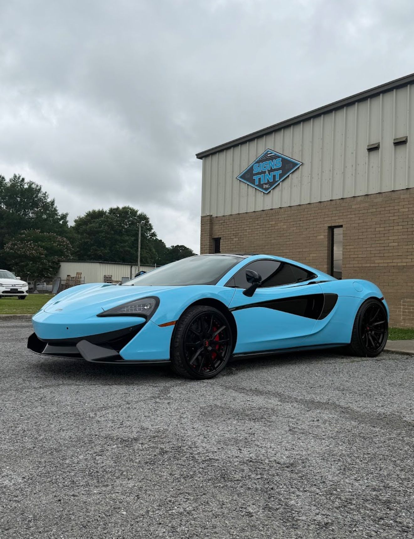 A light blue McLaren sports car parked on a gravel lot in front of a metal-sided building.