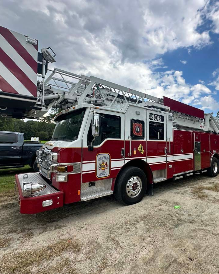 Red fire truck with ladder parked on a dirt lot under a partly cloudy sky.
