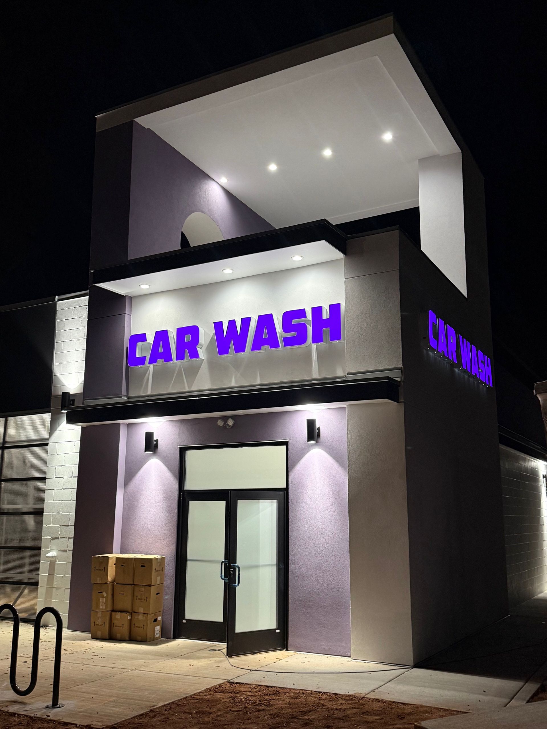 A brightly lit purple and white car wash building at night, featuring prominent purple signage above the main entrance.