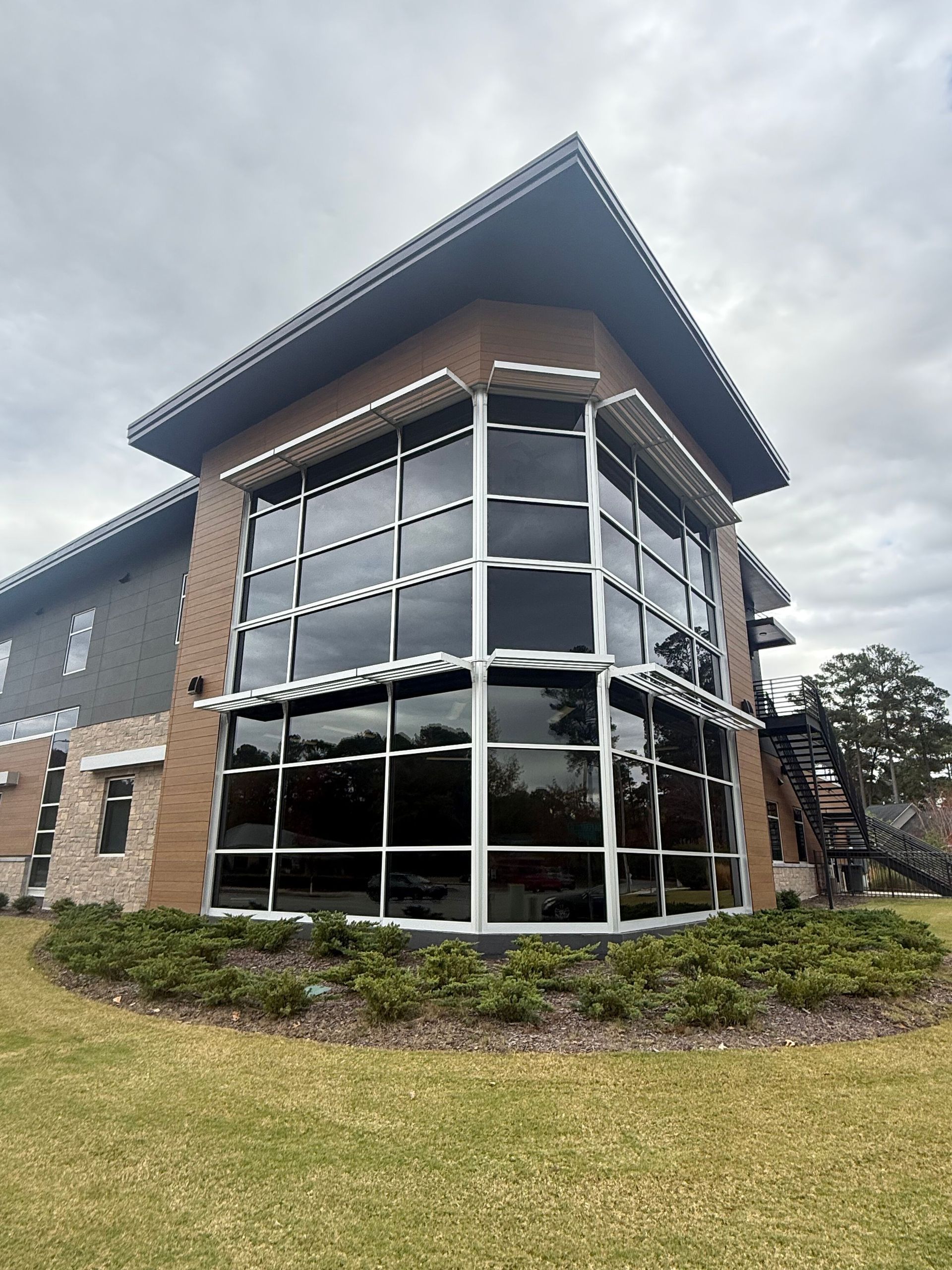A two-story commercial building with a large, curved glass window corner, brown siding, and dark trim under a cloudy sky.