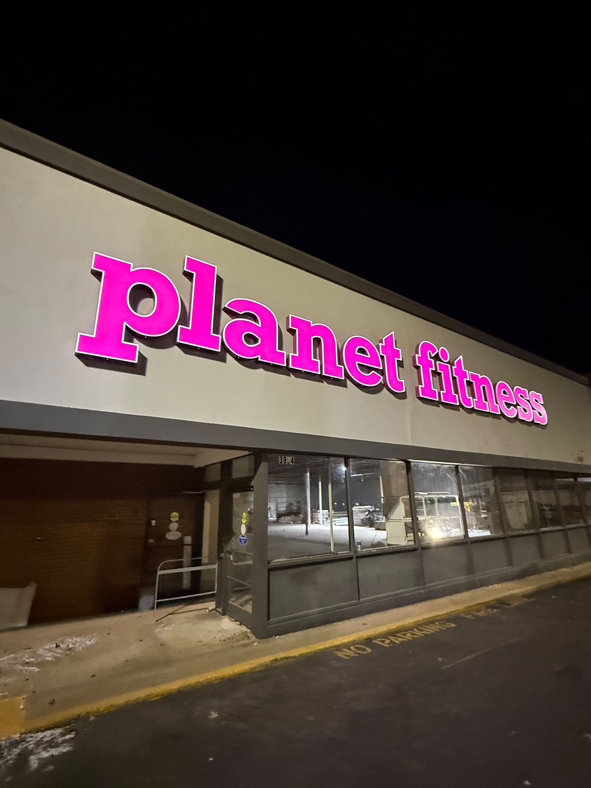 The exterior of a Planet Fitness gym at night, featuring a bright, glowing purple sign against a light-colored wall.