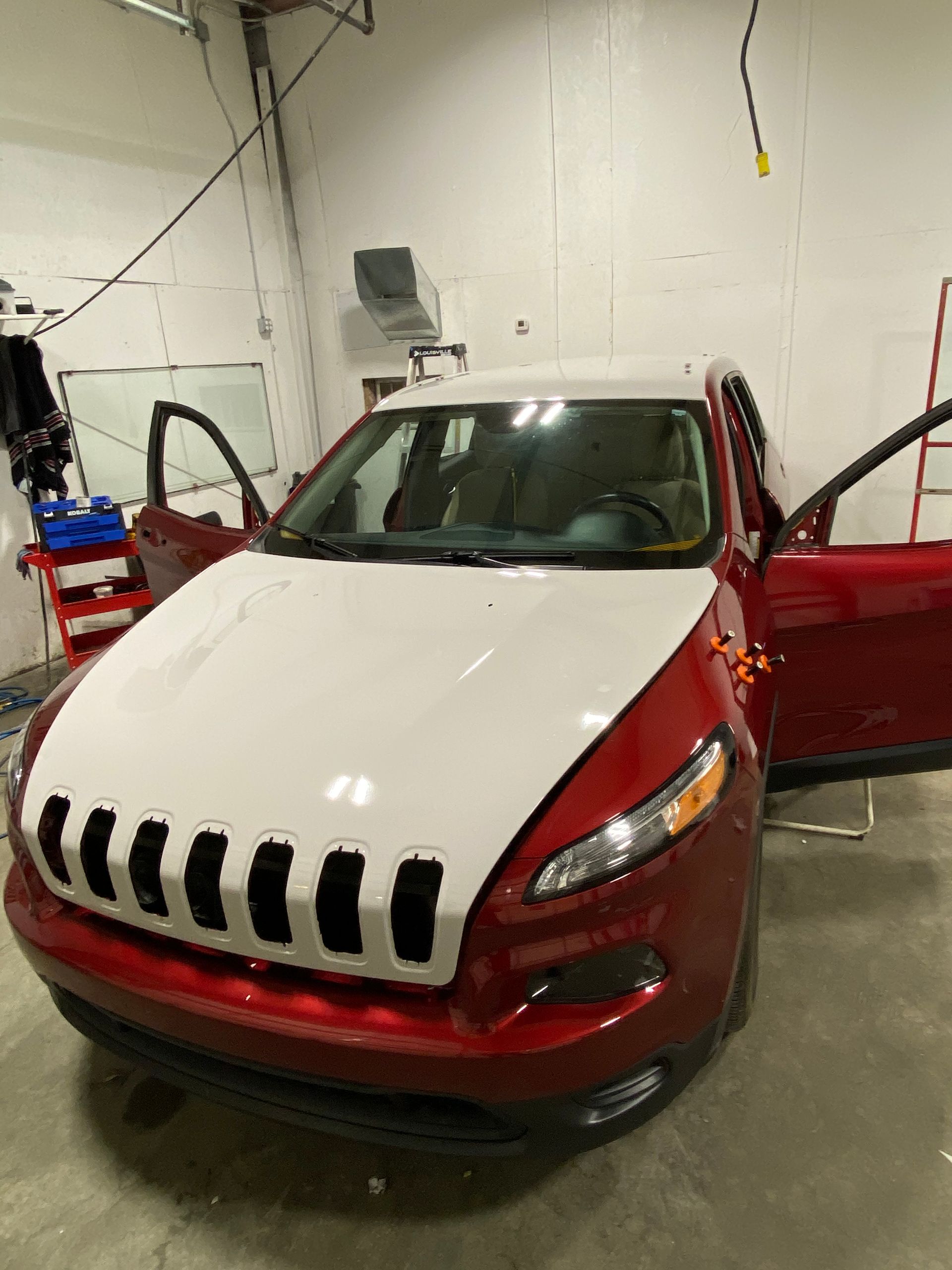 A red Jeep Cherokee with a white hood and roof parked inside a workshop with its doors open.