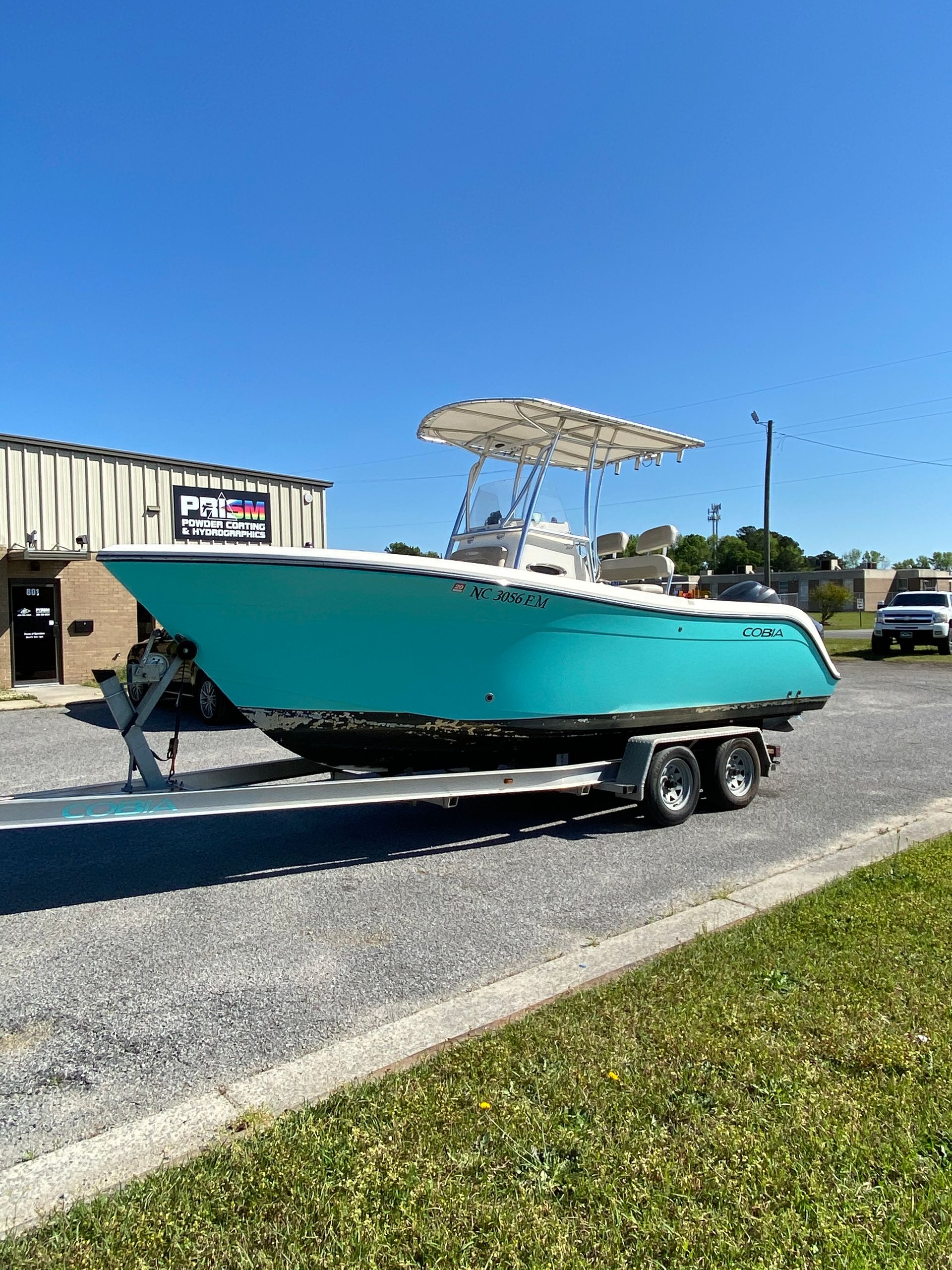 A turquoise center console boat on a double-axle trailer, parked on a gravel lot in front of a building under a blue sky.