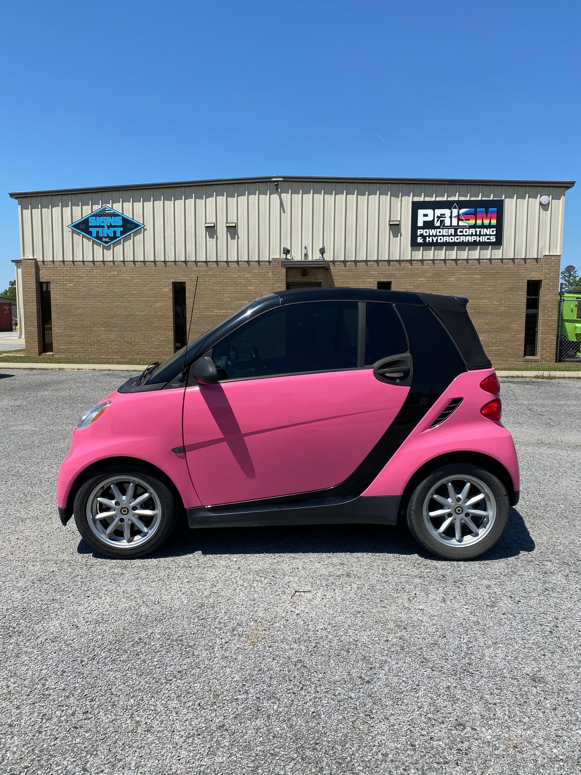 A bright pink Smart car parked on a gravel lot in front of a brick building with a Prism business sign.