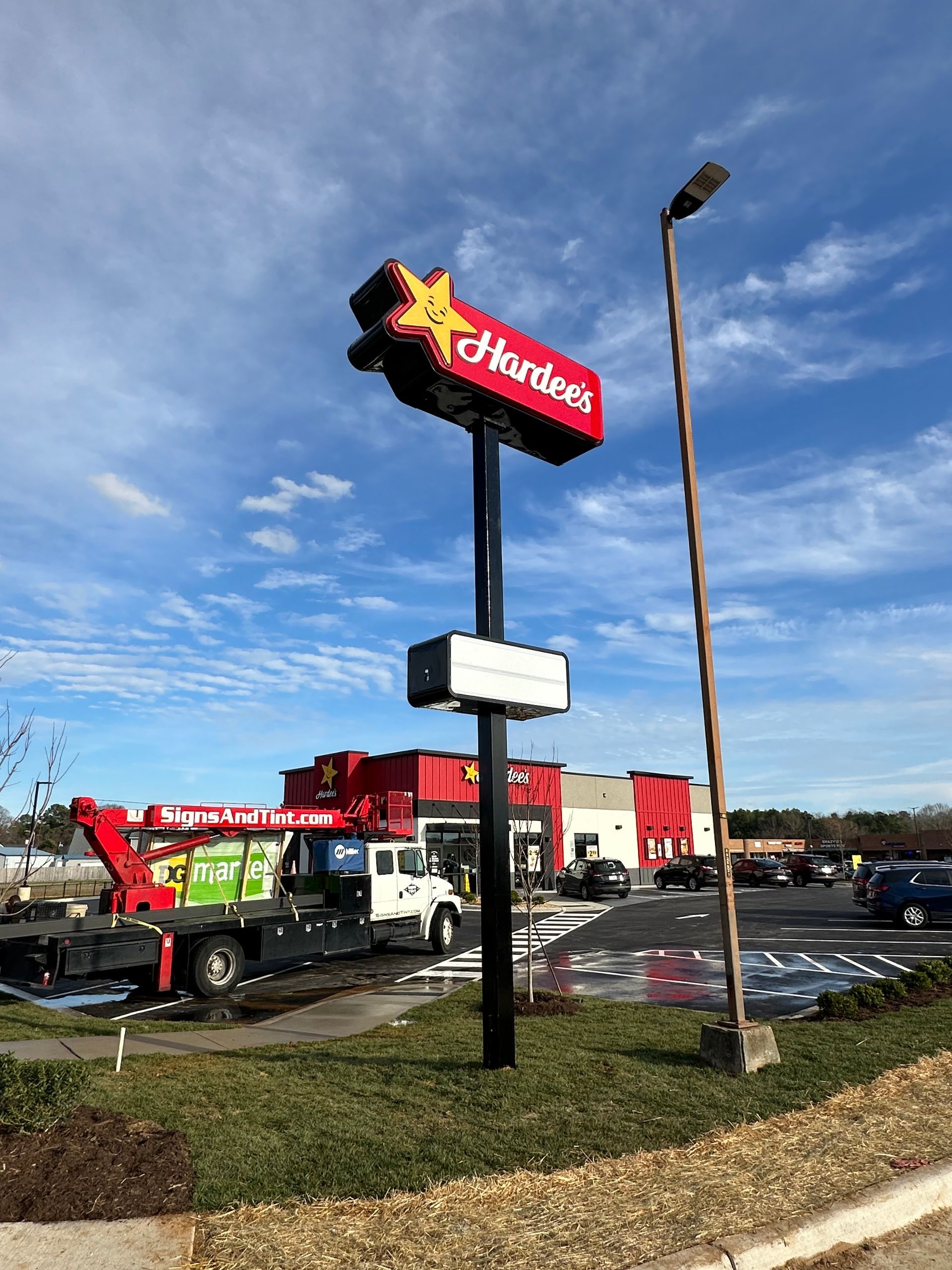 A red Hardee's sign stands by a utility truck in a restaurant parking lot under a blue, partly cloudy sky.