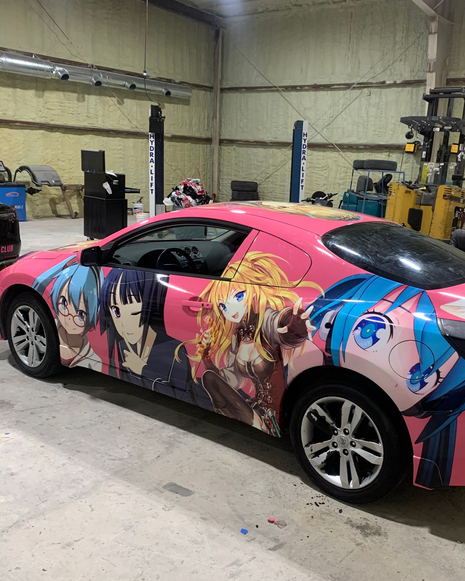 A pink car parked inside a workshop, featuring a full-body vinyl wrap with colorful anime characters along the side.