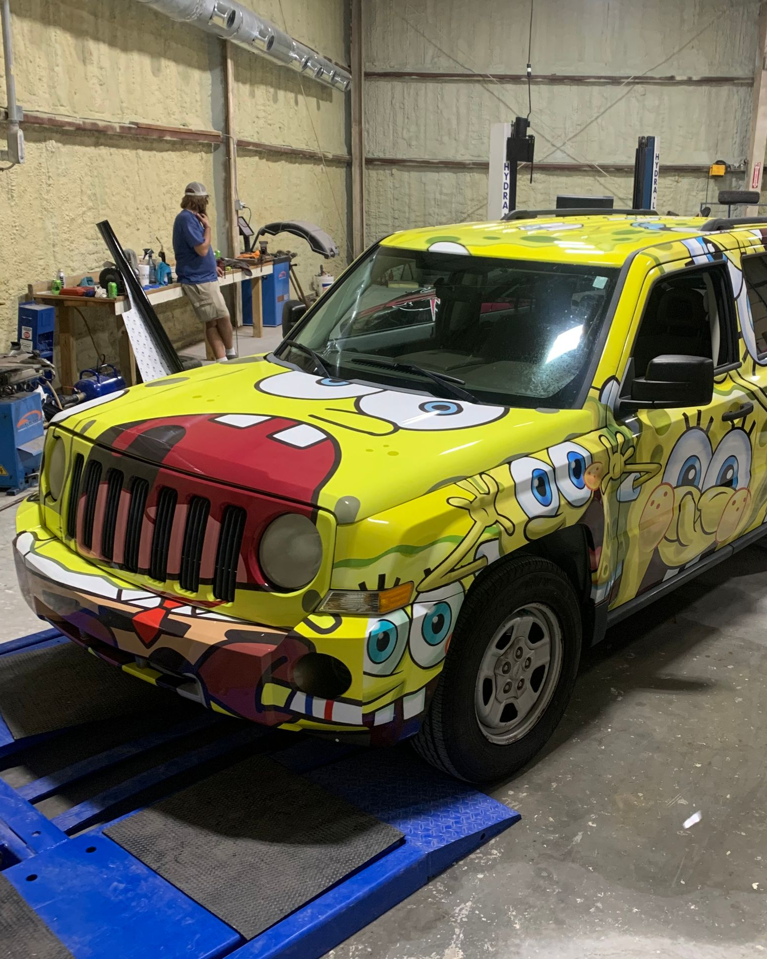 A bright yellow Jeep wrapped in a Spongebob Squarepants design sits on a vehicle lift inside an auto repair shop.