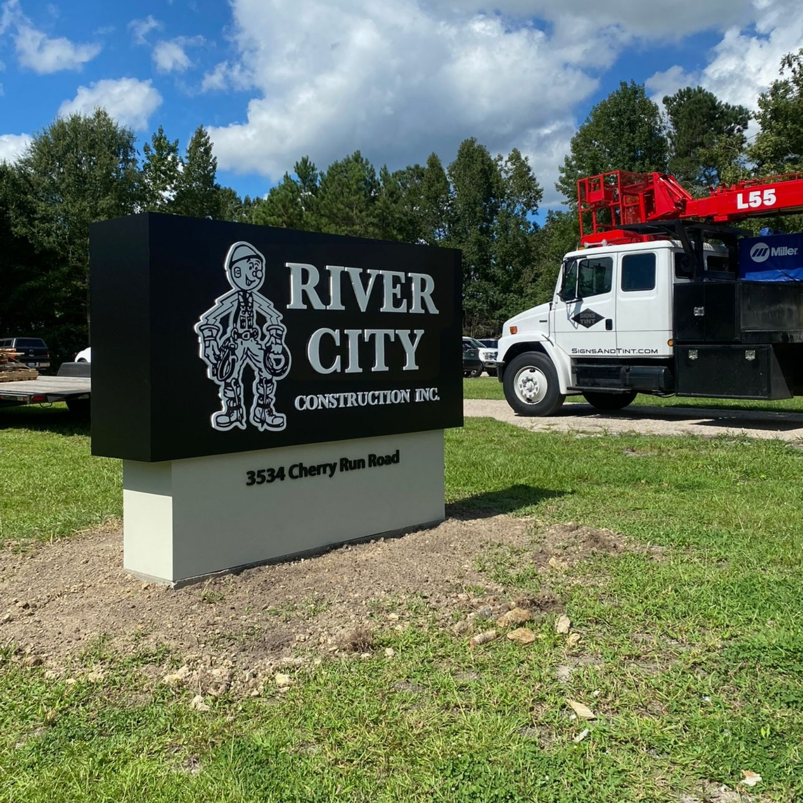 A black sign for River City Construction with a cartoon mascot, located outdoors next to a white service truck.