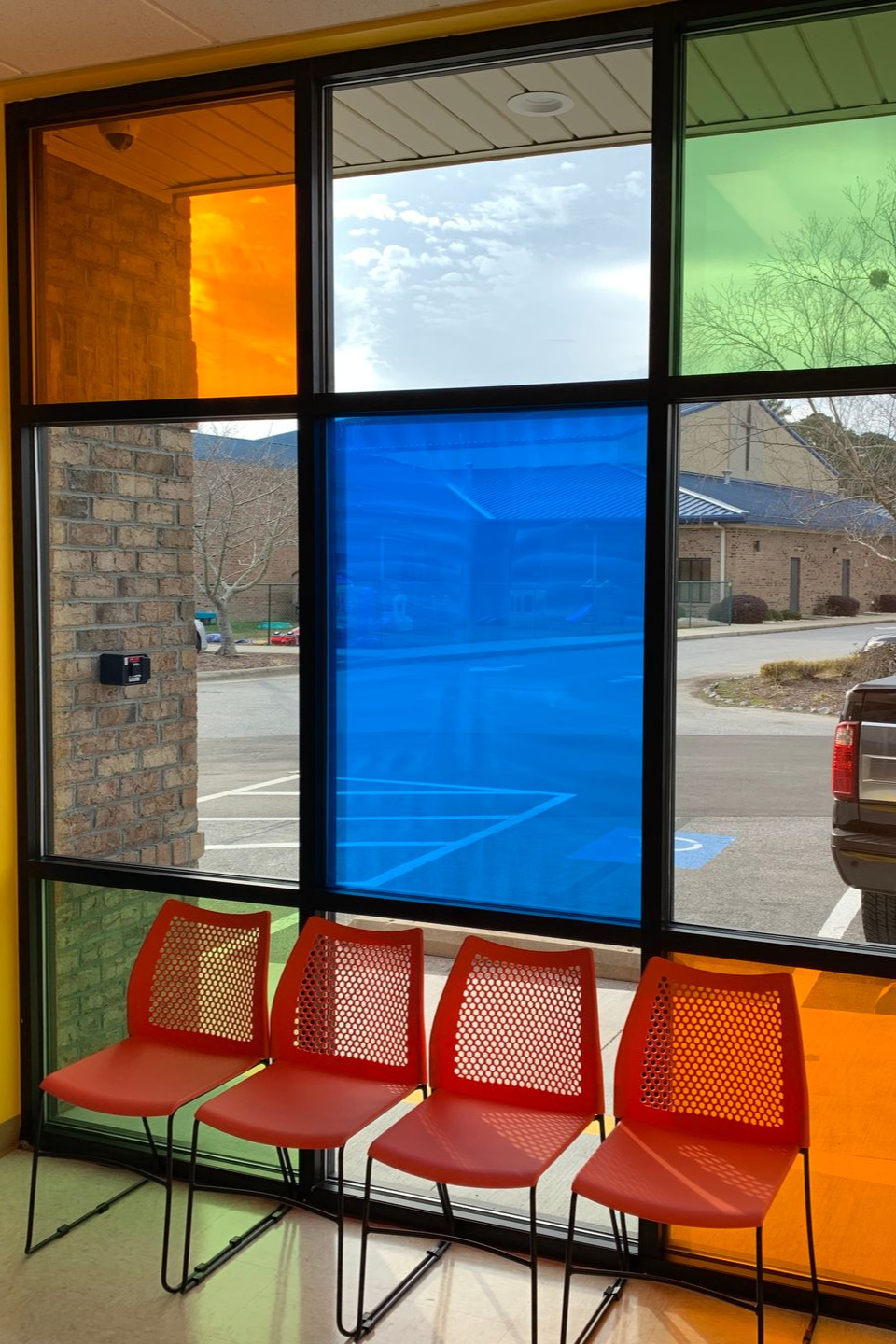Four orange chairs arranged in a row in front of a window with blue, orange, and green tinted glass panes.
