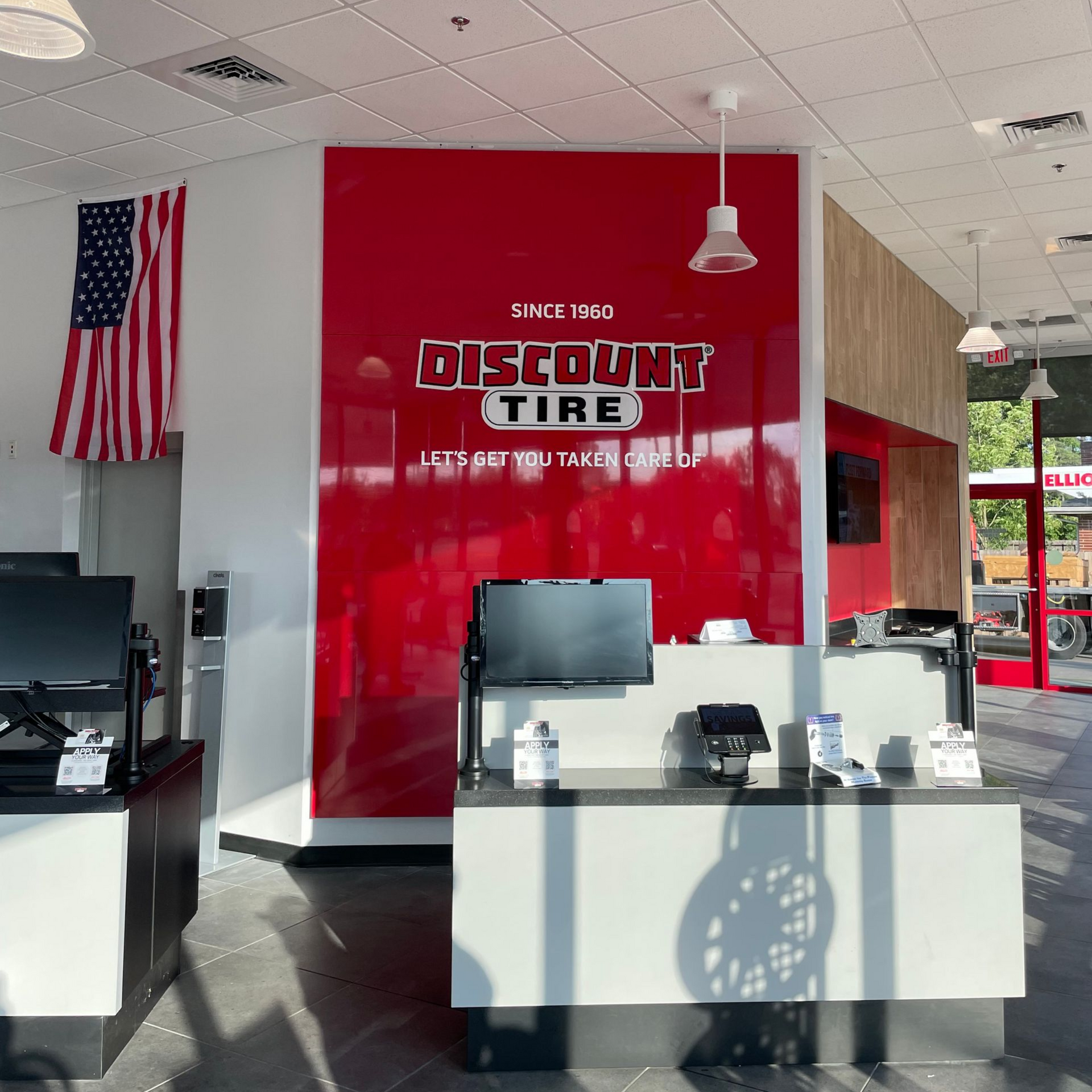 The interior of a Discount Tire store featuring a red accent wall with the brand logo, white service counters, and a flag.