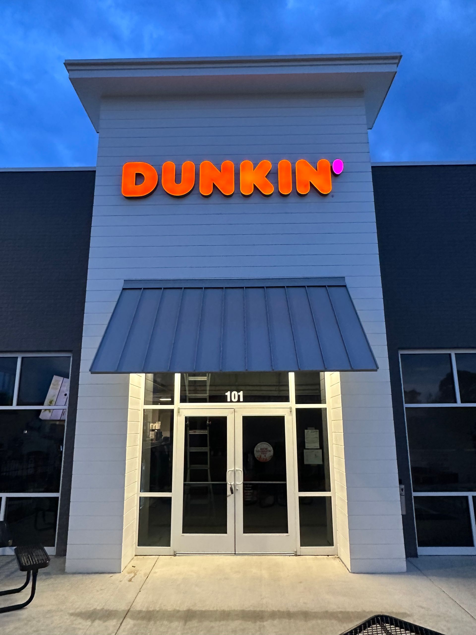 A Dunkin' storefront at twilight, featuring the orange brand logo on a white facade above glass entry doors.