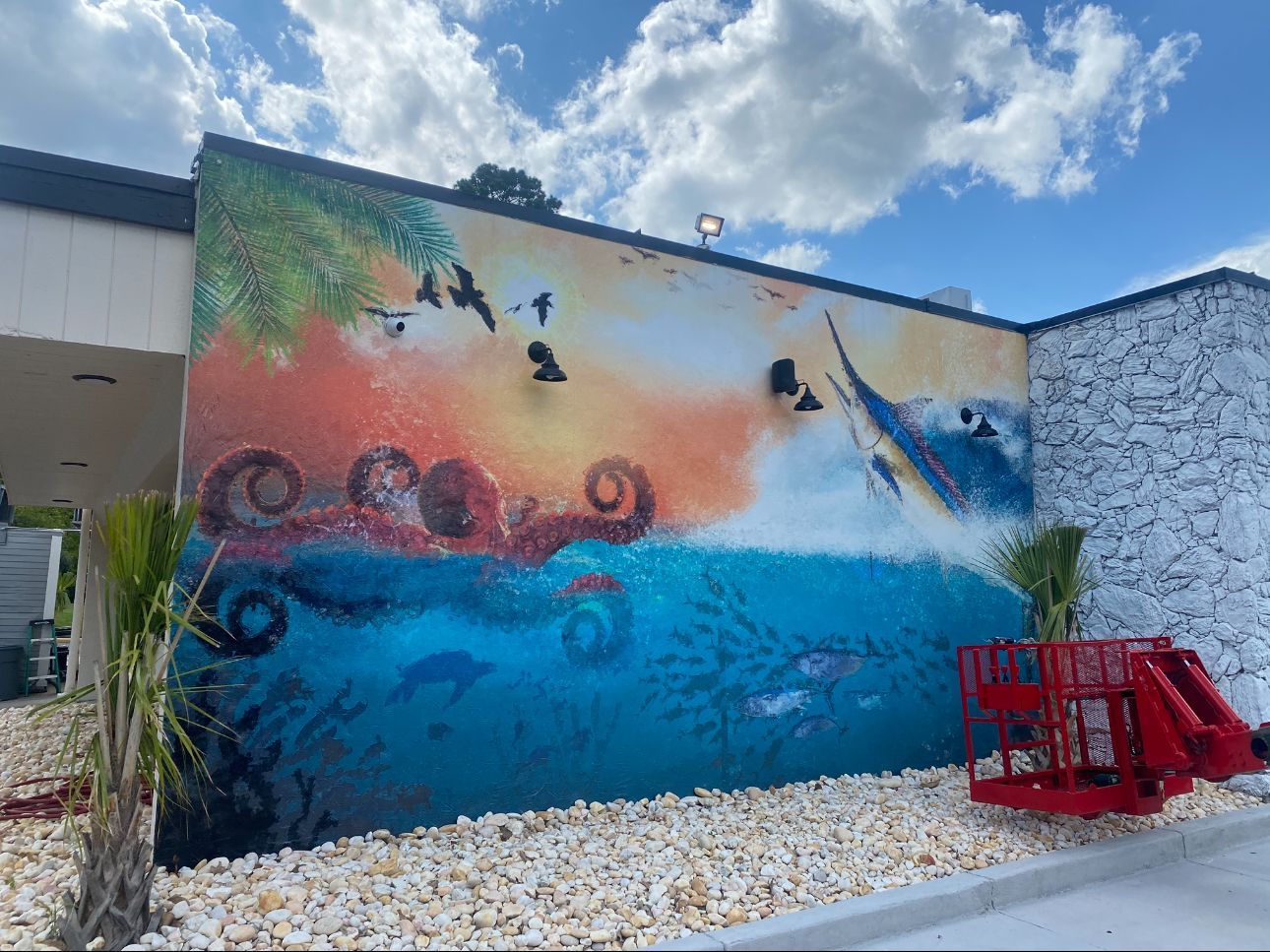 A vibrant mural on an exterior wall depicts a sunset, a leaping marlin, and an underwater scene with fish and coral.