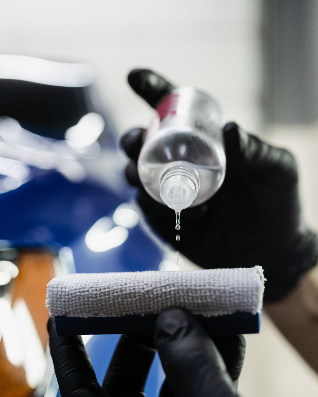 A person wearing black gloves drips a clear liquid from a bottle onto a detailing applicator sponge.