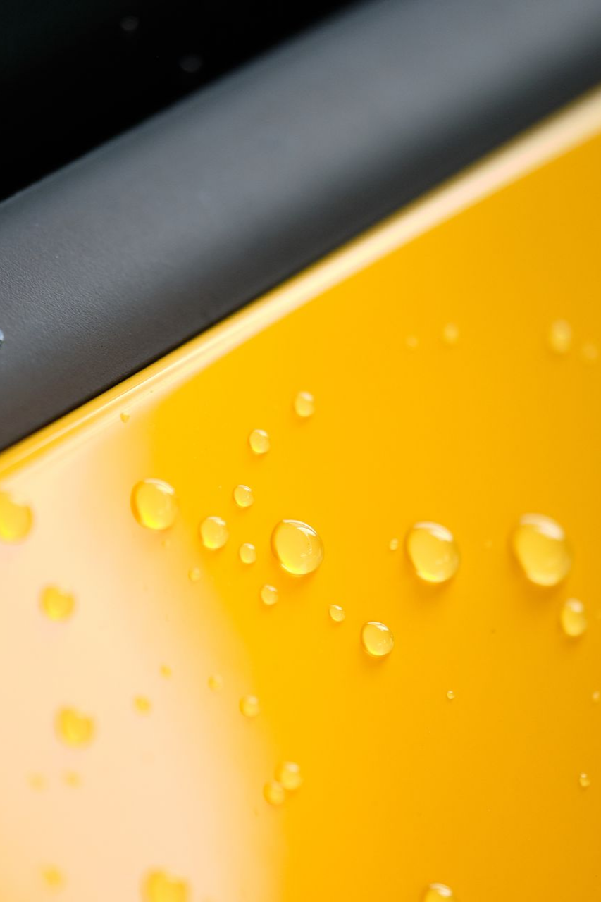 Close-up of clear water droplets resting on a smooth, glossy, bright yellow surface next to a black trim piece.