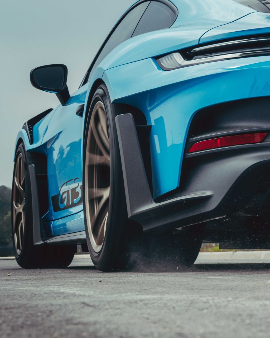 Low-angle shot of a bright blue Porsche 911 GT3 RS with bronze wheels and black aerodynamic trim on a paved surface.
