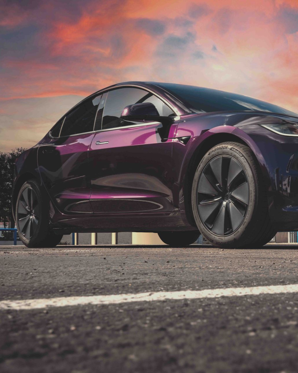 A purple Tesla parked on asphalt against a vibrant sunset sky.