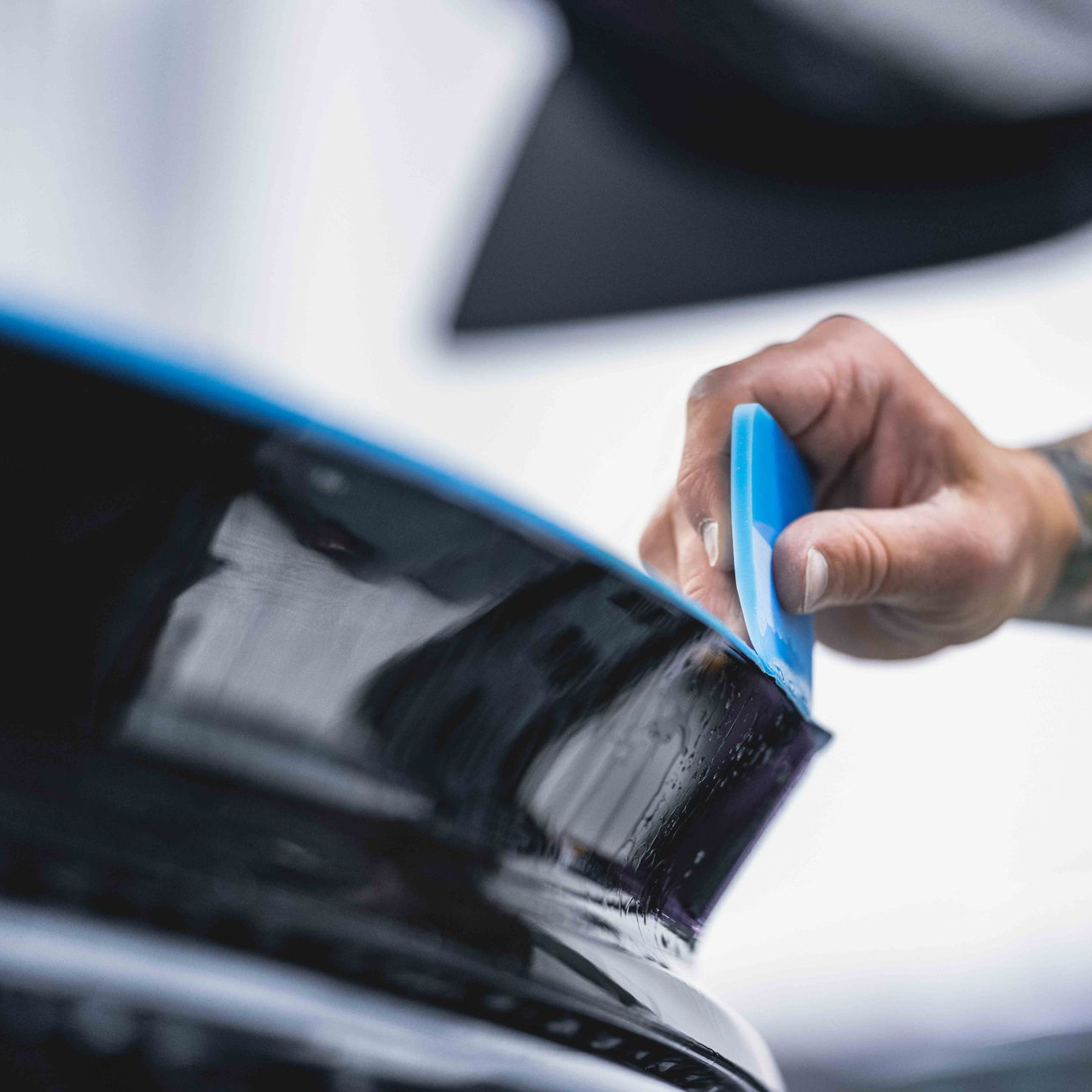A hand uses a blue plastic squeegee to smooth a protective film onto a glossy black car surface.