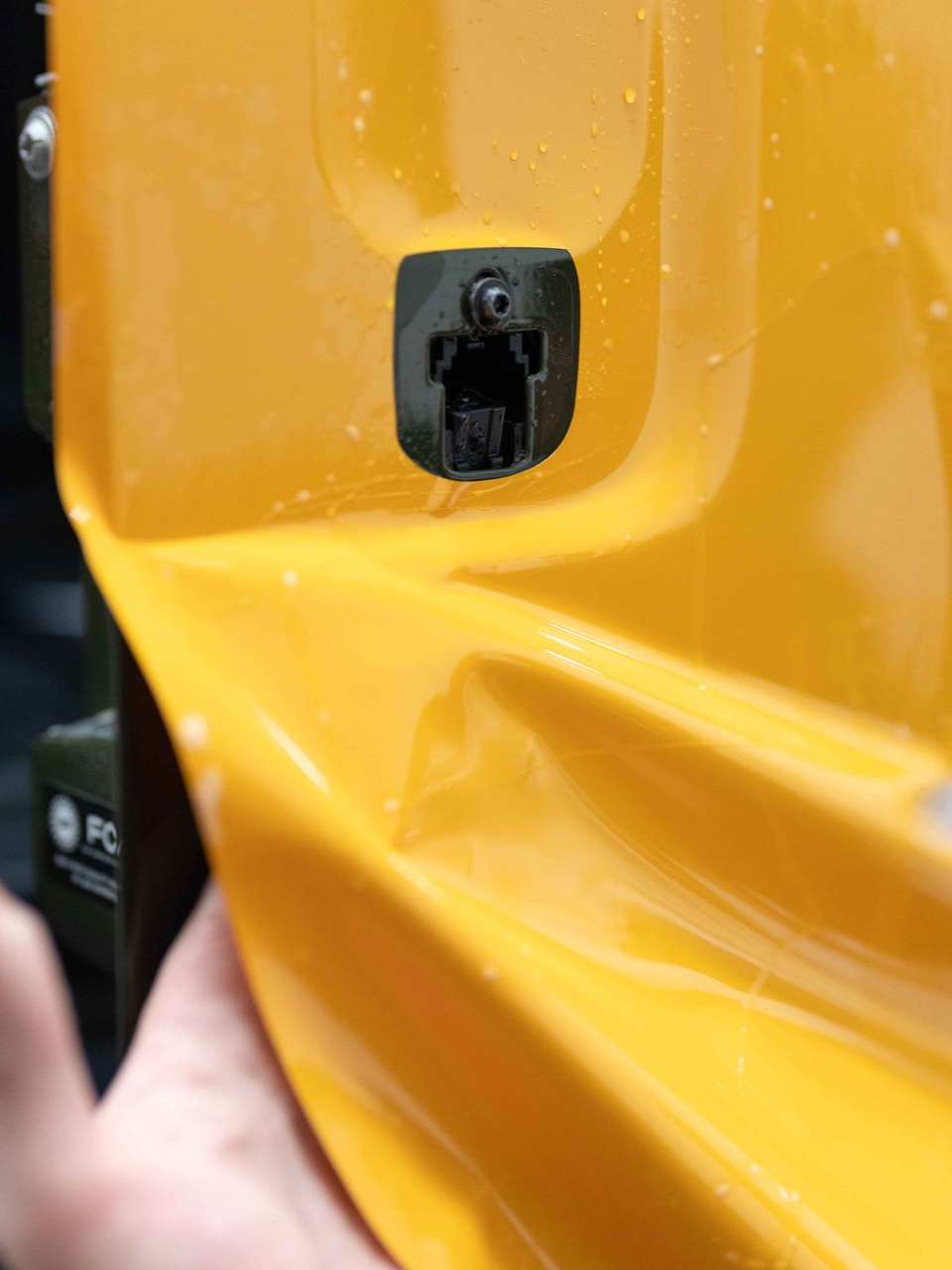 A hand holding a yellow vehicle body panel with a small black rectangular mounting point attached to its surface.