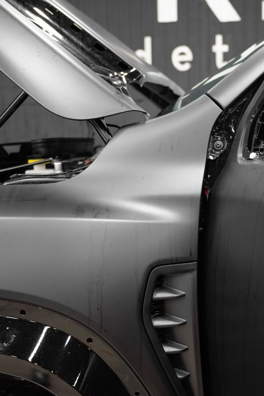 Side profile of a matte gray vehicle with an open hood, showing the fender, side vent, and front wheel arch.