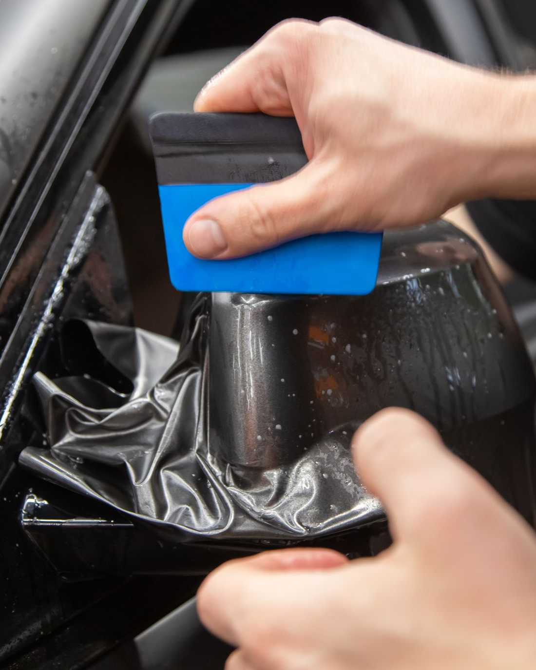 A person uses a blue squeegee to smooth black vinyl wrap onto a car's side mirror.
