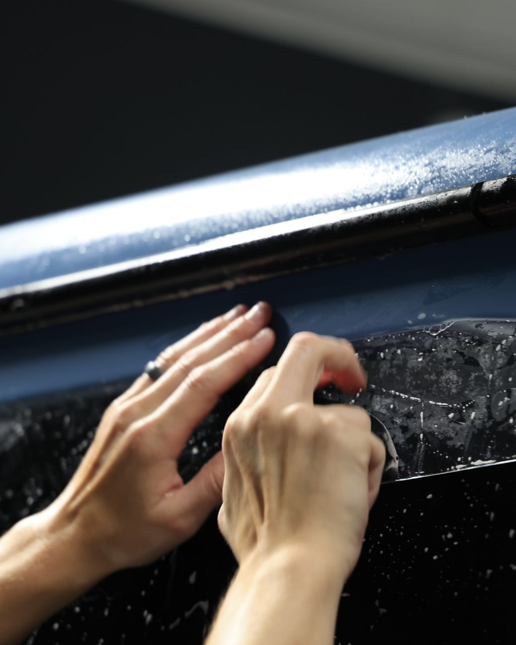 Hands applying dark, wet adhesive film to the edge of a blue car surface.