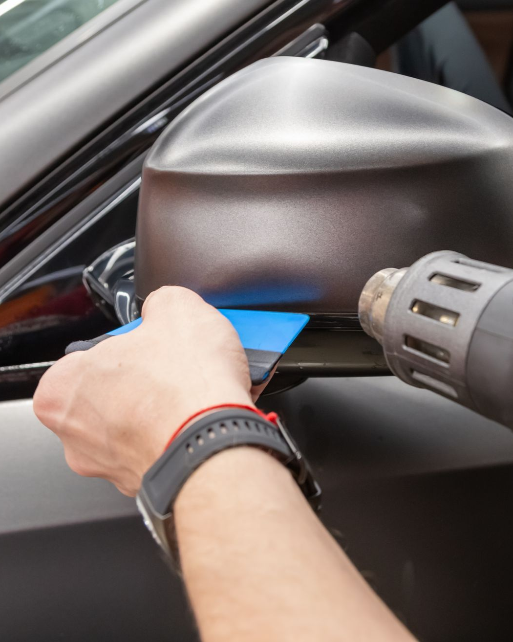 A person uses a heat gun and a blue squeegee to apply a vinyl wrap to a car’s side mirror.