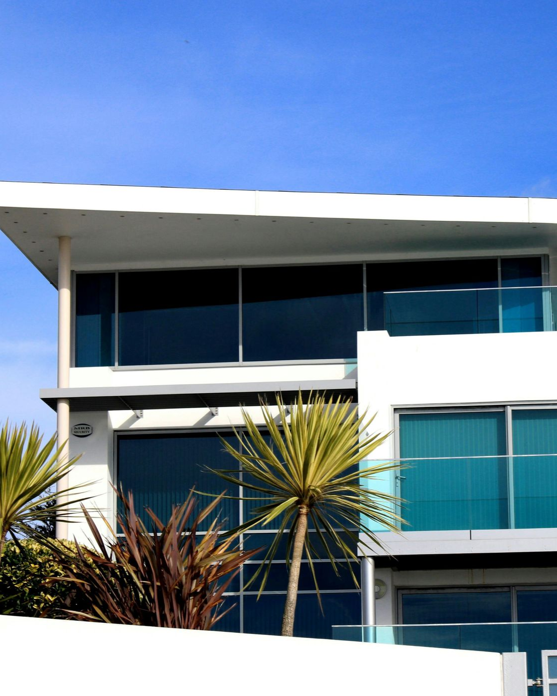 A modern multi-story home with white walls, large dark glass windows, and glass balconies, set against a blue sky.