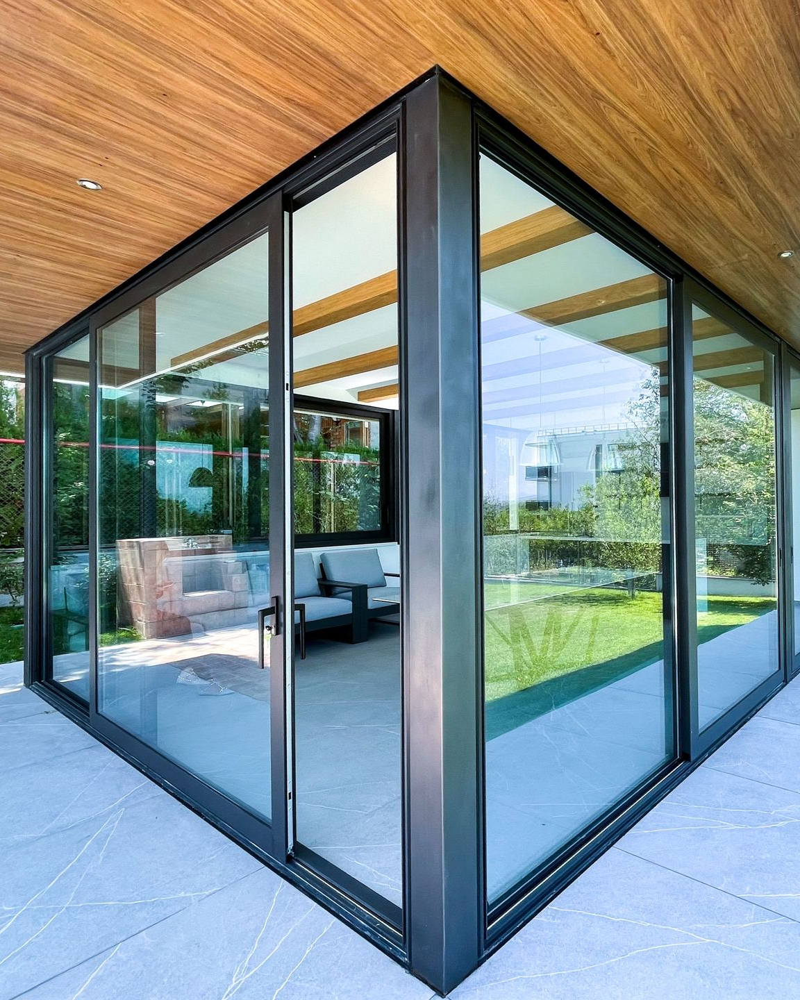 A modern patio with floor-to-ceiling glass walls, a dark metal frame, and a wooden ceiling overlooking a green garden.