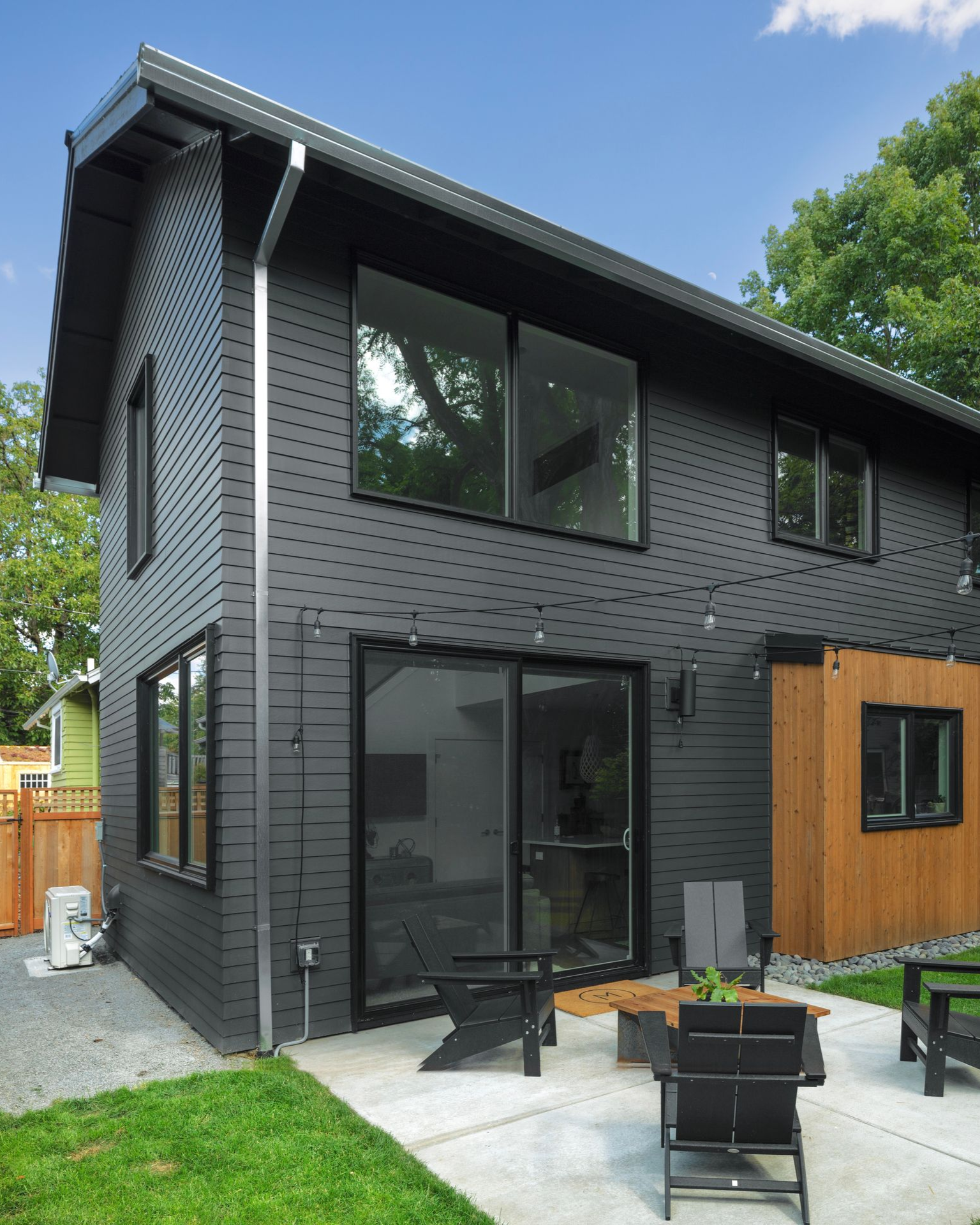 A modern two-story house with black horizontal siding, a metal downspout, and a patio with chairs in a backyard.
