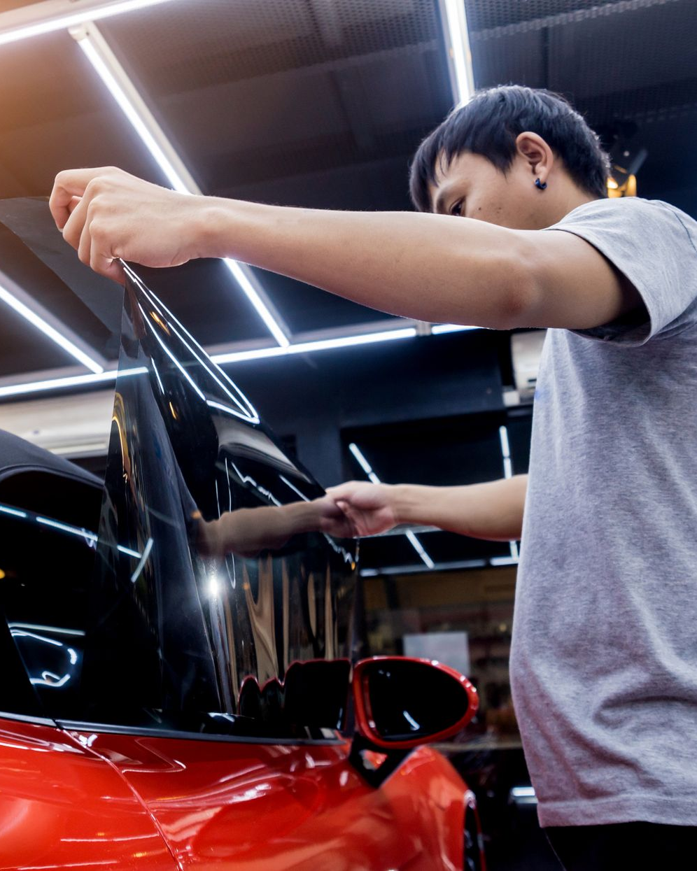 A person in a gray shirt carefully applies a dark window tint film to the side glass of a bright red car in a garage.