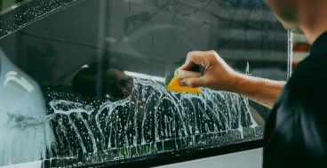 A person uses a yellow squeegee to remove soapy water from a glass surface, likely tinting a car window.