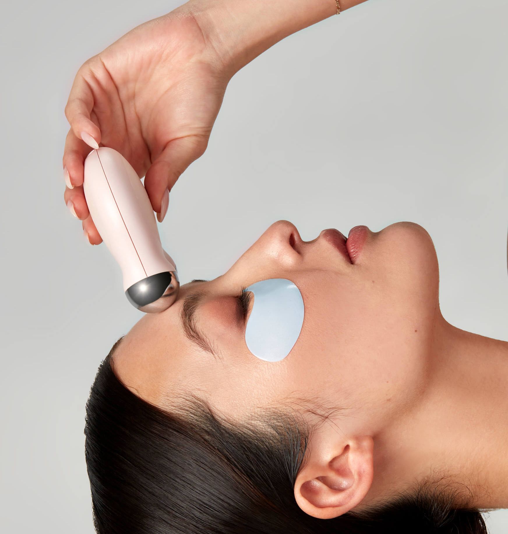 Woman using a handheld facial massager on her forehead with cooling eye pads on.