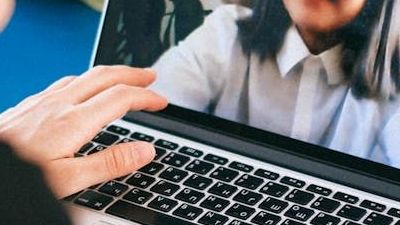 Hands typing on a laptop keyboard with a blurred person on a video call in the background