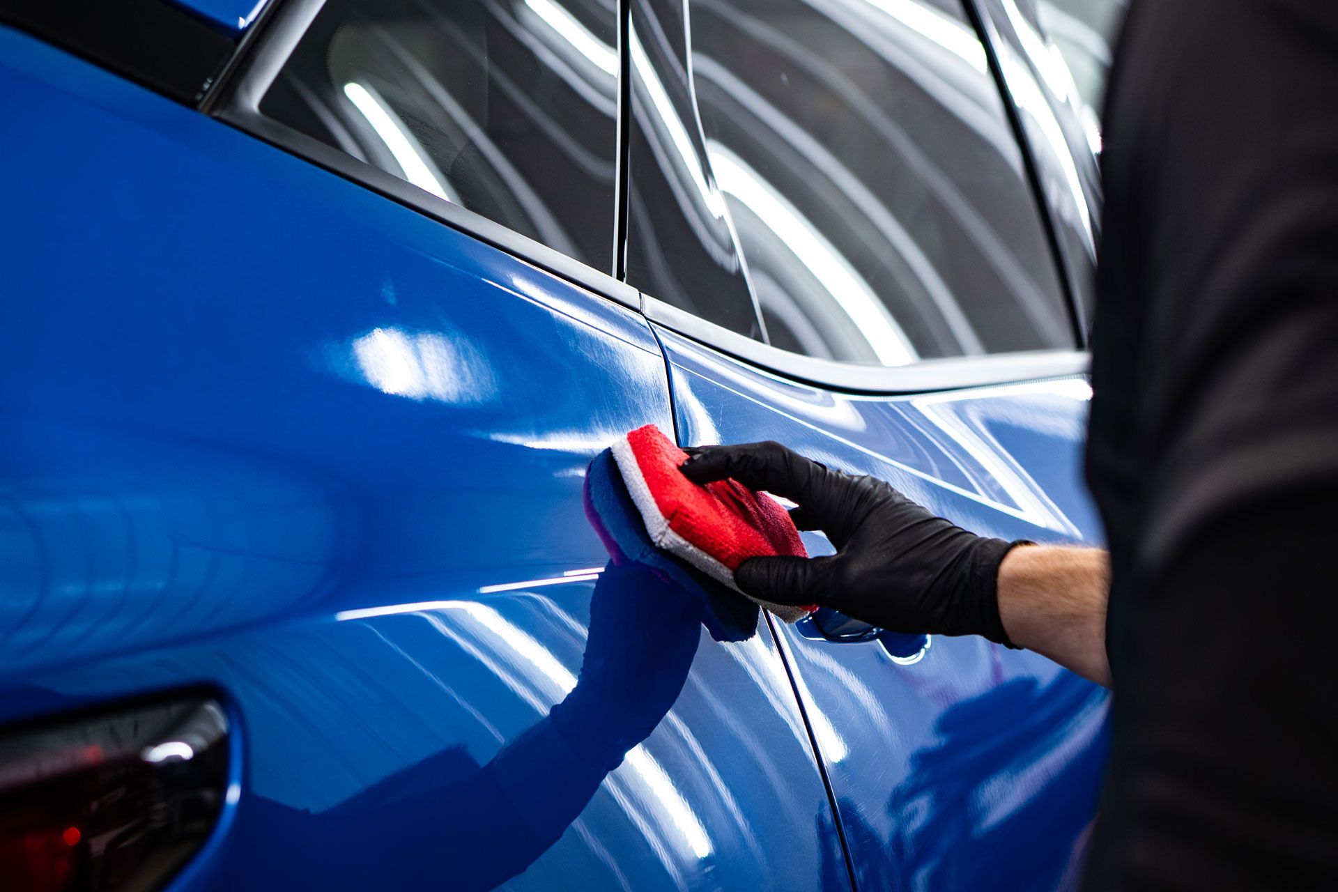 A person wearing a black glove uses a red applicator pad to apply ceramic coating to a glossy blue car door.