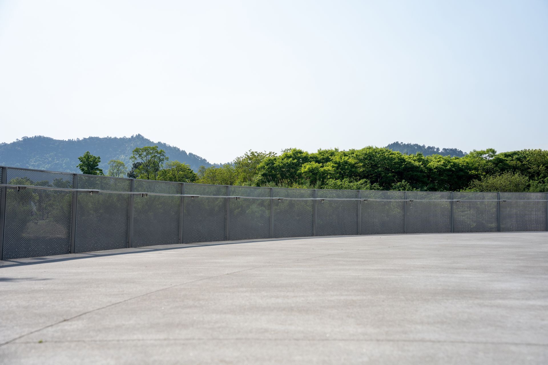 Empty concrete lot with chain-link fence, green trees, and distant hills under a clear sky